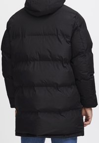 Mand iført en sort hættetrøje med puffet jakkel, vist fra ryggen, med hænderne synlige ved siderne og blå jeans synlige nederst under jakken.