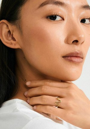Anillo ondulado de color dorado en una mano, con curvas suaves, sobre un fondo claro. La modelo lleva una blusa blanca.