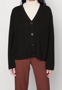 Cárdigan negro de punto con escote en V, tres botones grandes, puños y dobladillo acanalados, combinado con pantalones de tiro alto color óxido.