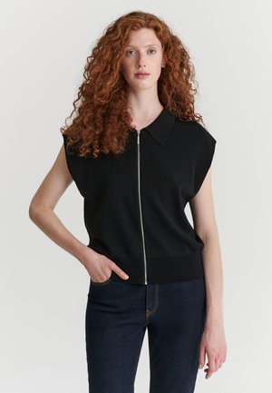 Jeune femme aux longs cheveux roux bouclés porte un haut noir sans manches avec fermeture éclair devant et un jean bleu foncé, debout avec une main dans la poche.