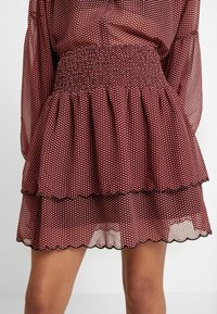 Burgundy lagerkjol med ett strukturerat nätmönster, som har en smockad midja och vågiga kanter. Lätt och genomskinligt tyg.