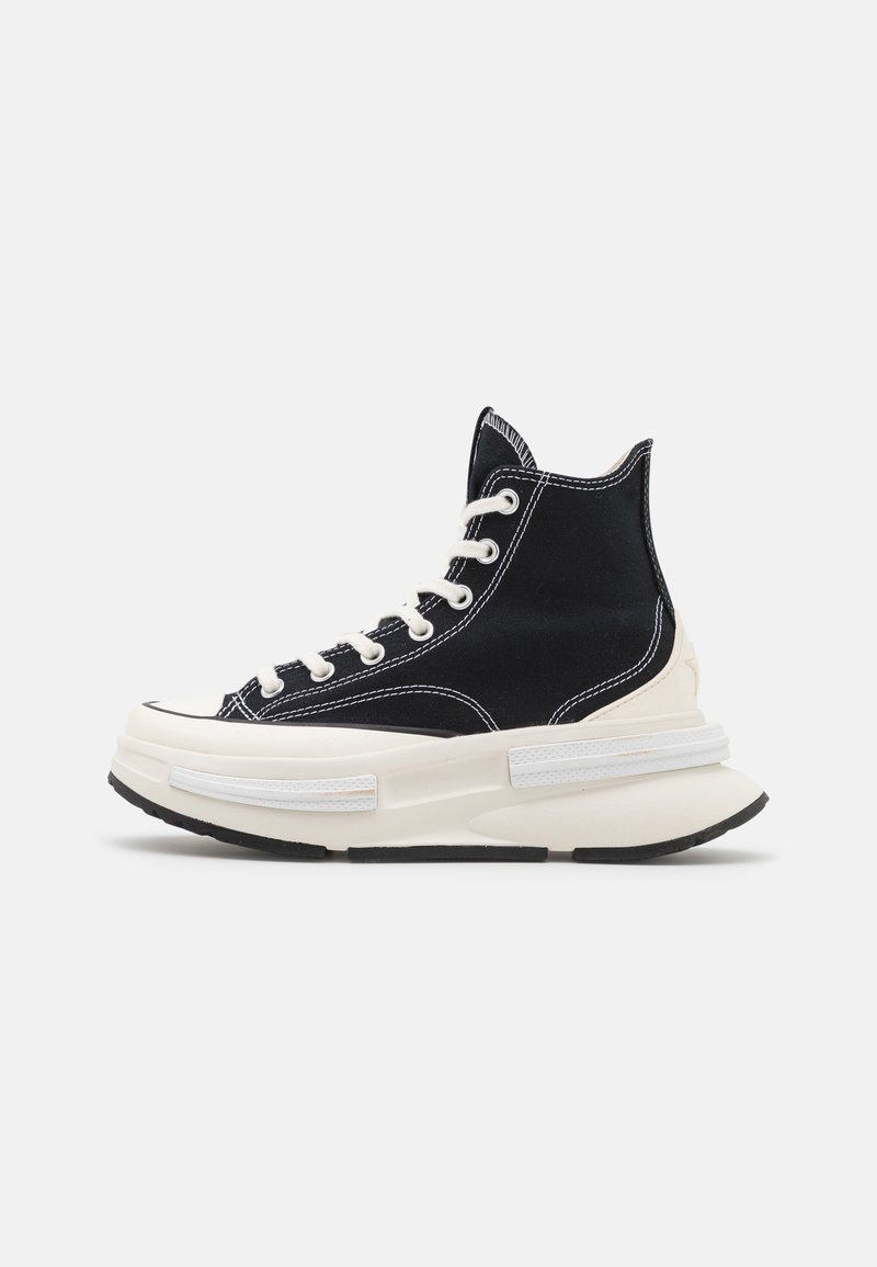 Basket montante noire avec couture blanche, lacets blancs, et semelle épaisse blanche avec semelle extérieure en caoutchouc noir sur fond uni.