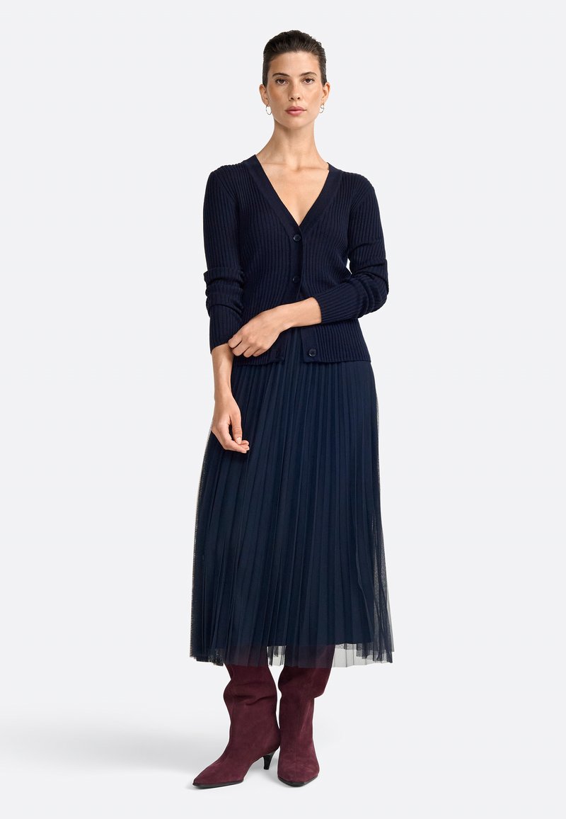 Cardigan navy a coste con scollatura a V e bottoni abbinato a una gonna plissettata in tulle navy. Stivali al ginocchio in suede bordeaux completano l'outfit.