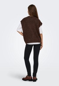 Femme debout de dos, portant un pull sans manches marron par-dessus une chemise blanche, un pantalon noir et des bottines marron à lacets sur un fond uni.