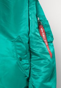 Teal, satin bomberjacka som visar en sidovy med en blixtlåsficka och en röd etikett märkt "TA BORT INNAN FLYGNING." Har en slät textur.