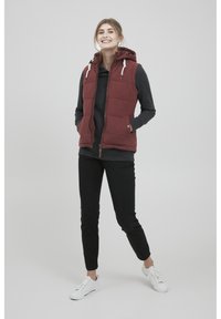 Gilet matelassé bordeaux avec capuche, fermeture éclair, poches avant et doublure grise, porté sur un haut sombre et associé à un pantalon noir et des chaussures blanches.