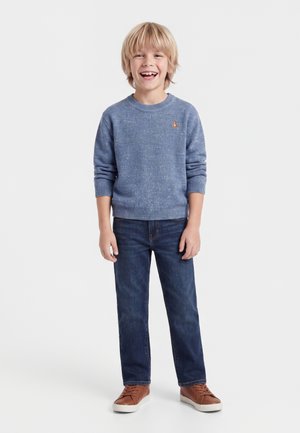 Garçon souriant aux cheveux blonds portant un pull bleu, un jean foncé, et des baskets marron, debout devant un fond blanc uni.
