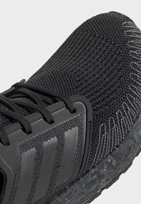Gros plan sur l'avant d'une chaussure de sport noire avec une tige en tissu tricoté, des lacets noirs, une semelle texturée et un motif gris subtil sur le côté.