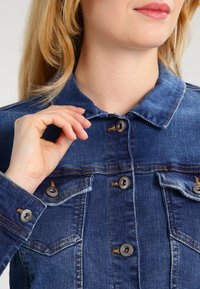 Dżinsowa kurtka z kołnierzykiem, pięcioma guzikami, dwoma kieszeniami na piersi, w ciemnoniebieskim kolorze i wyrazistym przeszyciem. Gładka tekstura.