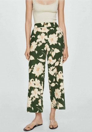 Persoon draagt een hooggetailleerde, wijduitlopende donkergroene broek met grote beige bloemenprint en een nudetanktop, gecombineerd met sandalen met doorzichtige bandjes.