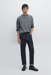 Grijze gebreide trui met een donkerblauwe kraag, gecombineerd met donkerdenim broek, opgerolde mouwen, en zwarte veterschoenen.