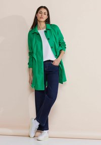 Grön, knäppt trenchcoat med en mjuk textur, lager på en vit skjorta. I kombination med mörka jeans och vita sneakers.