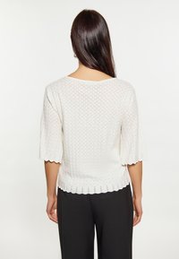 Top bianco a maglia con maniche corte e ondulate. Caratterizzato da un motivo a diamante testurizzato e un ampio scollatura. Indossato con pantaloni neri.