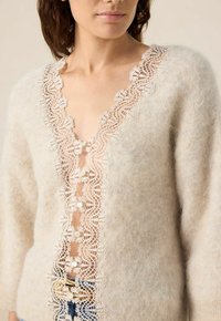 Pull en tricot beige avec un décolleté en V profond et une bordure en dentelle. Présente des boutons en perles le long de l'ouverture avant. Texture douce et duveteuse.