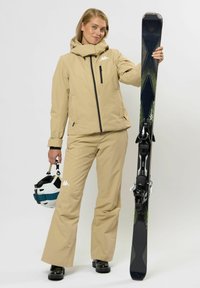 Frau in beigem Skianzug, die in einer Hand einen Ski und in der anderen einen weißen Helm mit blauer Sicherung hält, vor einem weißen Hintergrund steht.
