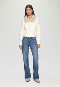 Weißer gerippter Crop-Pullover mit einem Kragen aus Kunstpelz, kombiniert mit blauen Schlagjeans und beigen Ankle-Boots. Einfaches Design, lässiger Look.