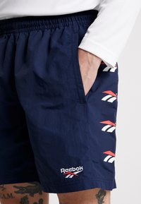 Námořnické modré šortky Reebok s elastickým pasem, s bílými a červenými akcenty a detailem loga, a bočními kapsami. Texturovaná tkanina.