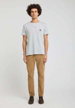 Jeune homme aux cheveux bouclés portant un t-shirt gris clair, un pantalon beige et des chaussures marron foncé, debout devant un fond clair uni.