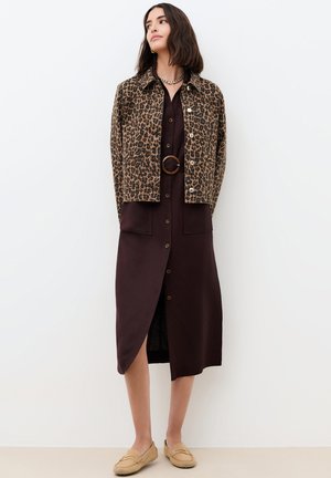 Mujer con chaqueta de estampado de leopardo sobre un vestido midi marrón abotonado con cinturón y mocasines color cuero, de pie frente a un fondo liso.