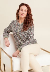 Bomberjacke mit Blumenmuster und grünem Saum, mit Reißverschluss, aus weichem Material. Kombiniert mit hellen, enganliegenden Hosen.