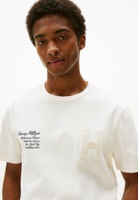 Camiseta de algodón blanca con un logo 'H' bordado y texto también bordado. Mangas cortas, escote redondo clásico, textura suave.