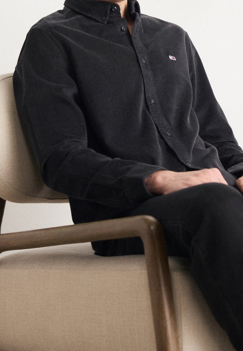 Camisa de pana negra con cuello abotonado. Presenta un pequeño logo, tejido texturizado y se combina con pantalones negros.