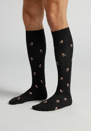 Sorte knæstrømper i knælængde med gentaget mønster af små peberkagefigurer iført røde hatte, lavet af blødt, strækbart stof.