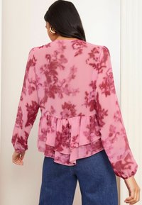 Langärmlige, pinke Bluse mit dunklem Blumenmuster, geschichteter Rüsche am Saum und weichem, transparentem Stoff. Getragen mit blauen Jeans, seitliche Ansicht von hinten.