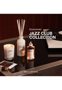 Une bougie allumée, un diffuseur à roseaux et un flacon vaporisateur de la collection Replica sur une table près d’un tourne-disque avec un texte sur la collection Jazz Club.