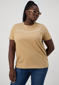 Žena s opletenými vlasmi, oblečená v béžovom tričku Tommy Hilfiger, modrých džínoch a oválnych béžových slnečných okuliaroch, ktorá sa pozerá nabok na jednoduchom pozadí.