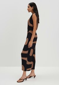 Mouwloze zwarte jurk met grote bruine abstracte vormen; heeft een elastische taille en reikt tot halverwege de kuit, gecombineerd met bruine hoge hakken.