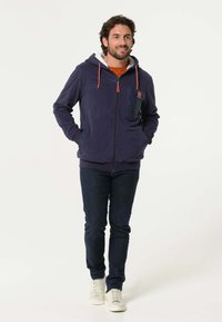 Marineblauwe hoodie met oranje koordjes, voorvak, ritsluiting en fleecelinning; gecombineerd met donkere jeans en witte sneakers.