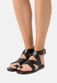 Černé kožené sandály s křížovými pásky, otevřenou špičkou a kotníkovým páskem se zlatou sponou. Plná podešev a hladká textura.