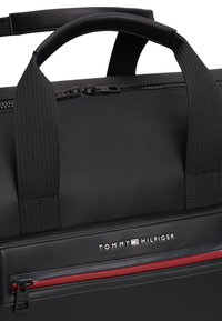 Μαύρη υφασμάτινη τσάντα με υφή στην επιφάνεια, με επάνω λαβή, κόκκινο φερμουάρ και πλακέτα με την επωνυμία "TOMMY HILFIGER" σε ασημί γράμματα.