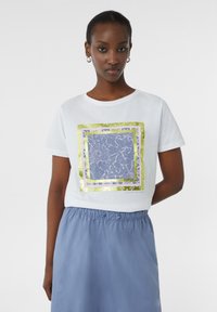 Witte katoenen t-shirt met een bloemig ontwerp in blauw en gouden folie-lijst; gecombineerd met een lichtblauwe rok met een trekkoord in de taille.