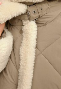 Chaqueta acolchada beige con un suave cuello y forro de borrego color crema, y detalles de cierre con hebilla metálica y broche.
