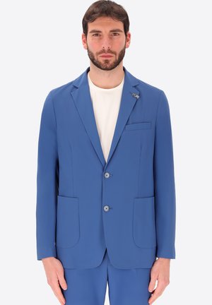 Uomo che indossa una giacca da completo blu sopra una camicia bianca, in piedi davanti a uno sfondo chiaro e uniforme, rivolto verso l'osservatore con un'espressione neutra.