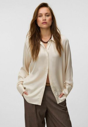 Femme aux longs cheveux bruns porte une chemise en satin crème boutonnée, un pantalon marron et un collier sombre de perles, les mains dans les poches.