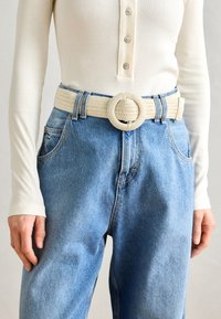 Wovenes cremefarbenes Band mit einer runden Schnalle, getragen über hellblauen hochtaillierten Jeans, ergänzt durch ein geripptes, langärmeliges weißes Oberteil.