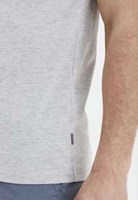 Un tricou gri din bumbac care arată o primăvară detaliată a cusăturii laterale, având un etichetă cu dungi alb-negru și o textură netedă și moale.