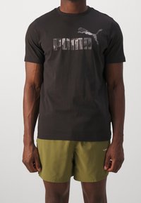 Černé bavlněné tričko s grafikou maskáčového loga PUMA. V kombinaci s olivově zelenými šortkami, které mají jednoduchý, sportovní design.