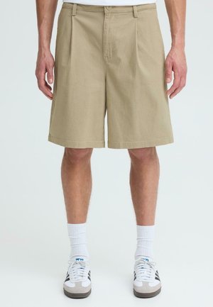 Mann iført beige knelangte plisserte shorts, hvite ankelsokker og hvite joggesko med svarte striper på ensfarget bakgrunn.