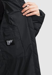 Quiltet sort ydertøj, der har en glat tekstur, et syet mærke og en åben front, der viser et kontrasterende inderskud.