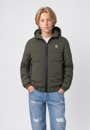 Teenager mit blondem Haar trägt eine dunkelgrüne gesteppte Kapuzenjacke und hellblaue zerrissene Jeans und steht mit den Händen in den Taschen.