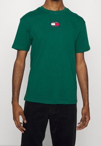 Man i en grön kortärmad T-shirt med en liten Tommy Jeans-logotyp på bröstet och svarta byxor.