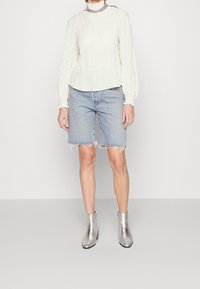 Weißes, strukturiertes Langarmoberteil, blaue Denim-Shorts mit ausgefranstem Saum und silberne Stiefeletten. Das Outfit weist ein geschichtetes Ausschnittdetail auf.