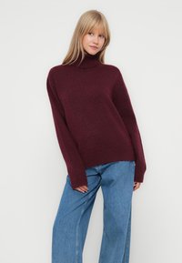 Pulover burgundy cu guler înalt, cu o croială lejeră, manșete și tiv cu textură. Asortat cu blugi oversize de culoare albastru deschis, creând un aspect relaxat.