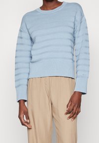 Maglione a righe blu chiaro in maglia a costine, con maniche lunghe e scollatura rotonda. Abbinato a pantaloni beige, che evidenziano un fitting rilassato.