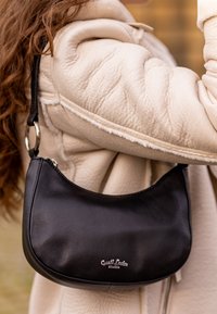 Černá kožená taška přes rameno s křivkovým designem, vybavená stříbrným kováním a prošitými detaily, vedle krémové prošívané bundy.