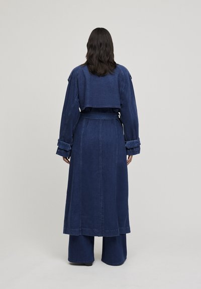 Cappotto lungo in denim di colore blu scuro con design a giacca corta, maniche ampie, dettaglio della cintura e pantaloni a gamba larga in abbinamento. Texture liscia.